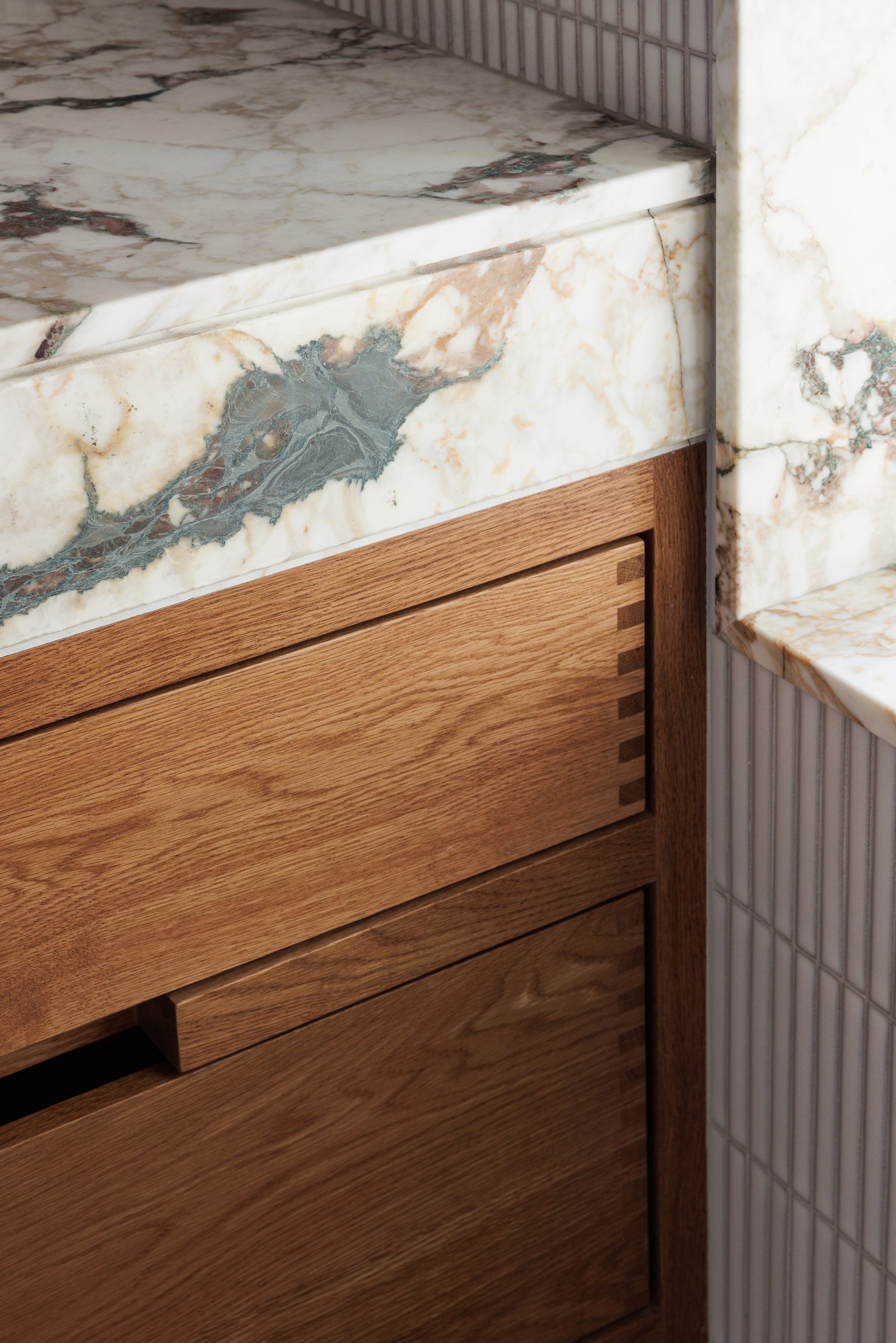 Right: a junction of veined marble and dovetailed oak, fluted tile turning the corner behind.
