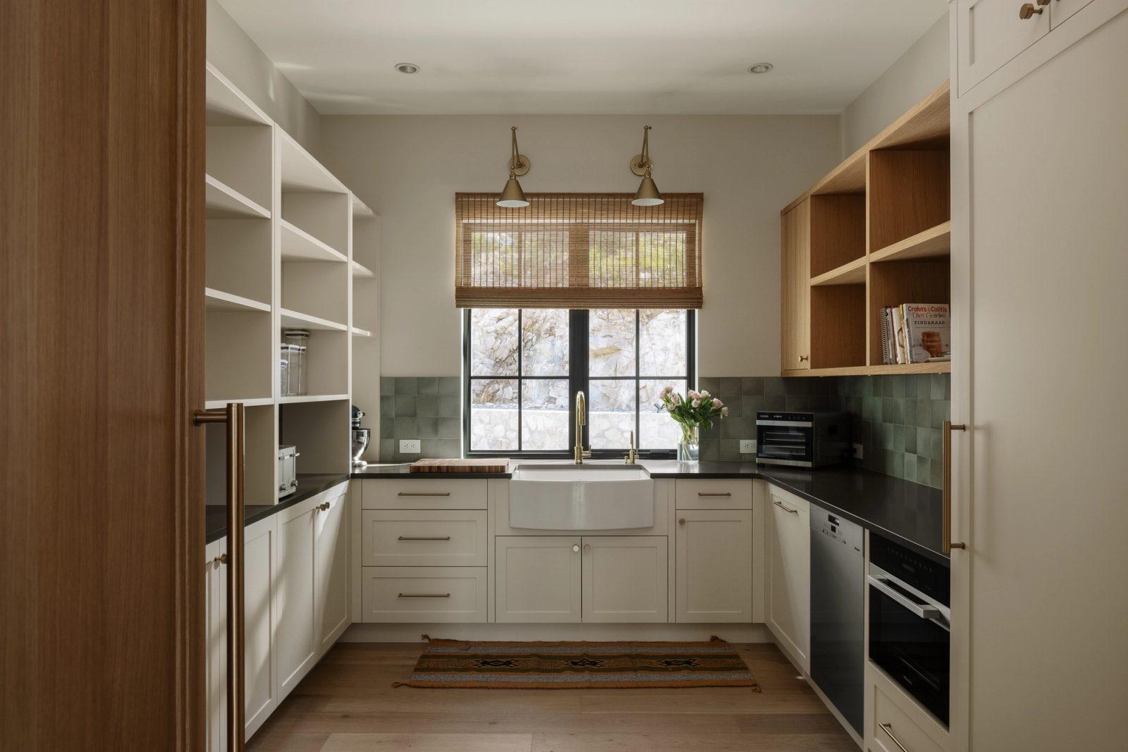 A working pantry in soft green tile and dark counters, a farmhouse sink beneath a window filled entirely by cut rock.