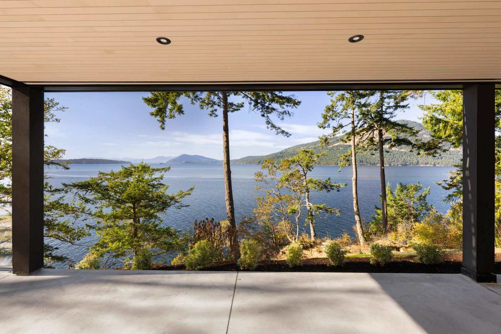 The view out from beneath a deep overhang: standing firs, the channel, and a forested island ridge across it, with no part of the house in frame.