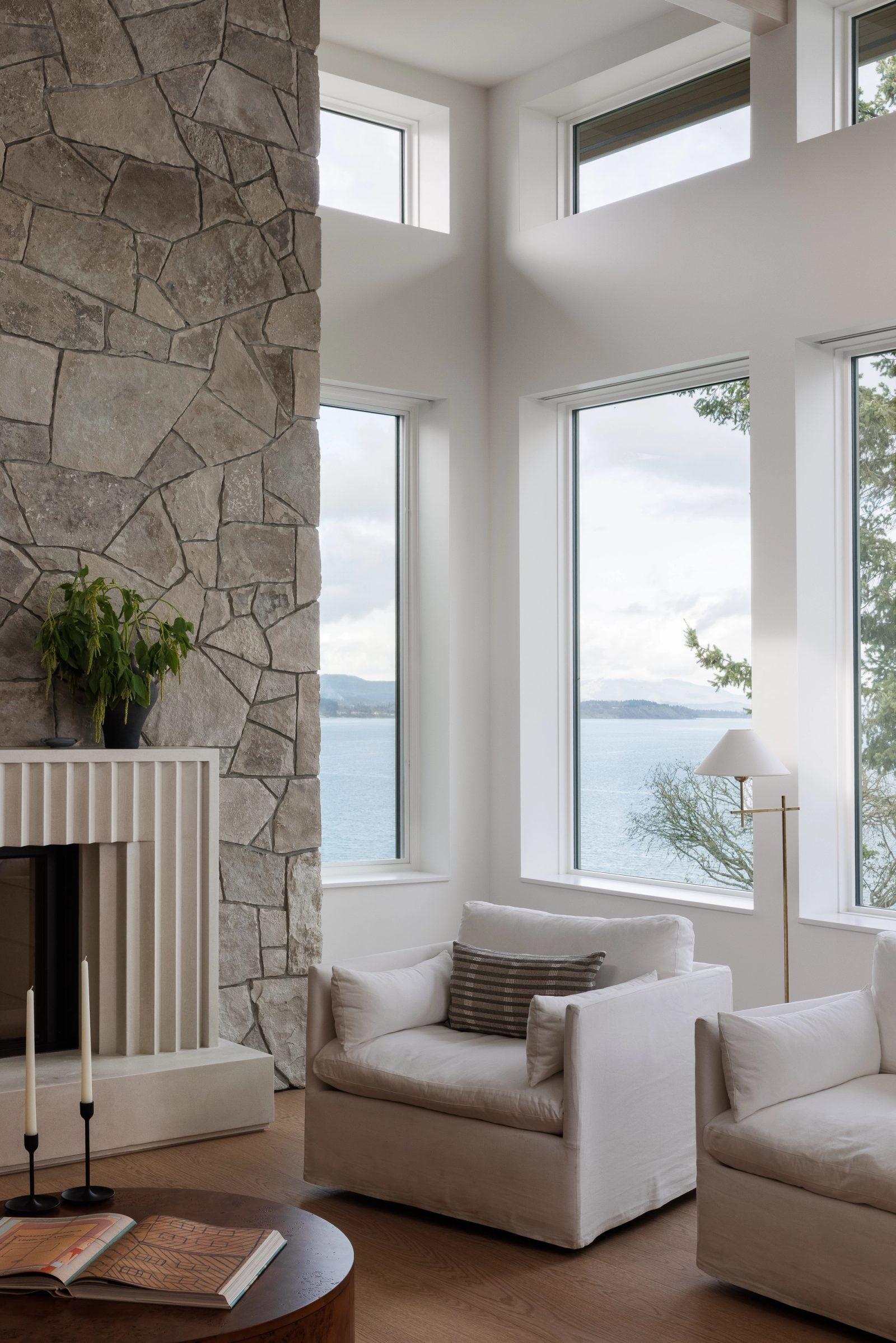 Left: a living room corner glazed on two sides above the water, a stone chimney rising beside it and a fluted cast-stone fireplace at its foot.