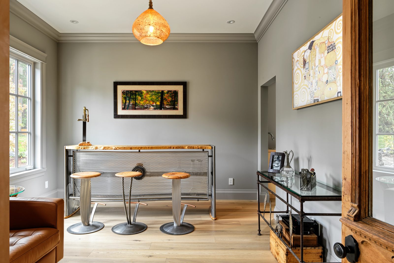 Sitting room in warm grey with a curved steel bar under a live-edge wood counter, three metal stools and an amber glass pendant overhead.