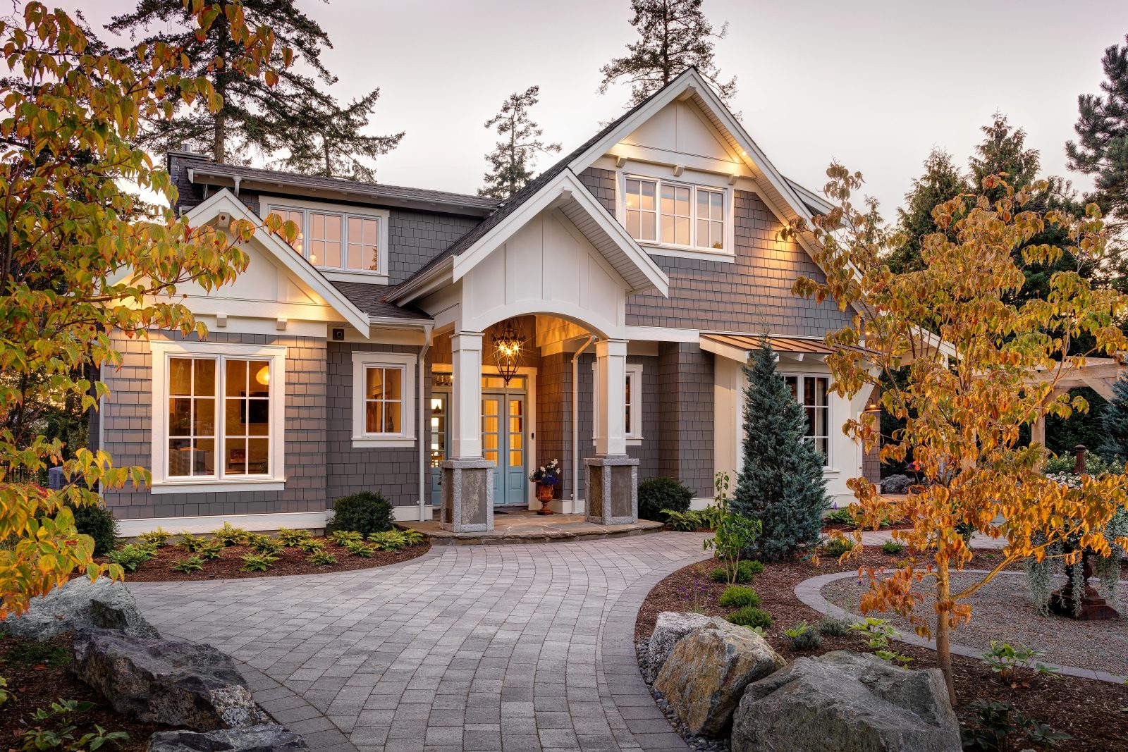 Shingled entry porch lit from within at dusk, framed by dogwood branches in autumn colour, with a paver walk curving between granite-based columns.