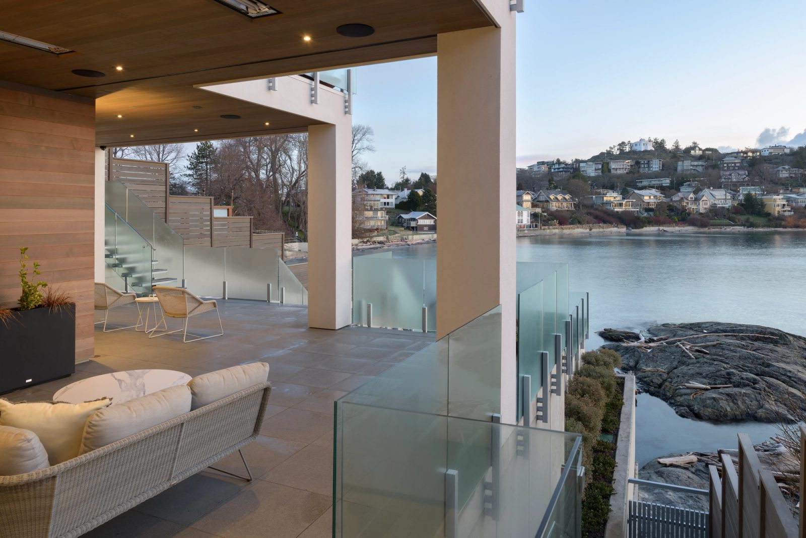 Covered terrace at dusk looking past a glass guard to rock, driftwood and calm water, with houses on the far shore.
