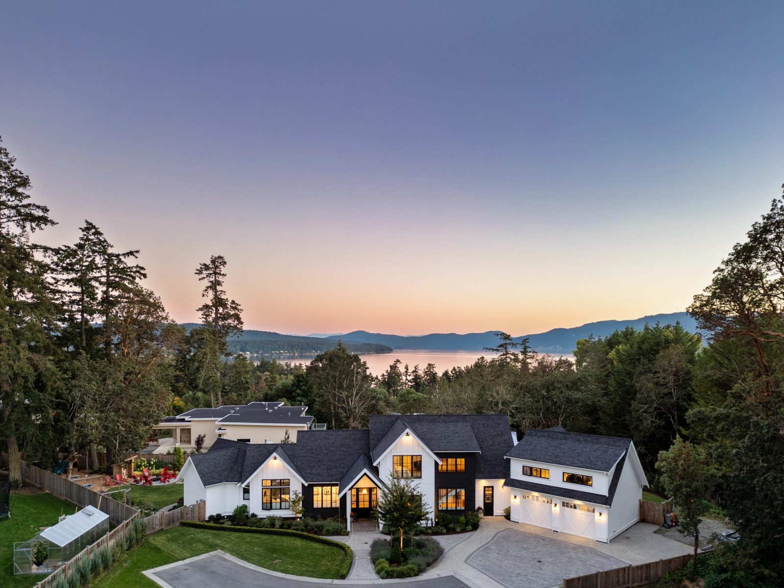 Aerial view at dusk of a modern farmhouse with a detached garage, lawn and greenhouse, with the inlet and hills beyond