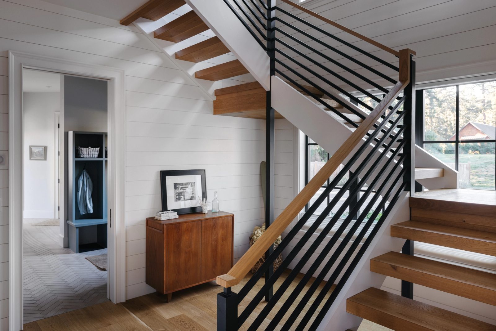 Open-riser oak stair with a black metal balustrade, with a herringbone-tiled mudroom and blue lockers beyond