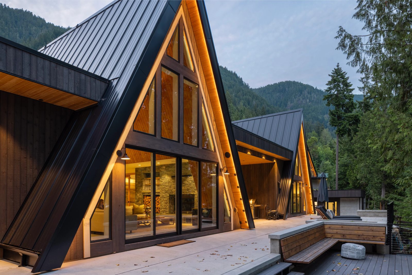 Deck at dusk with a stone fireplace glowing behind glazed A-frame walls