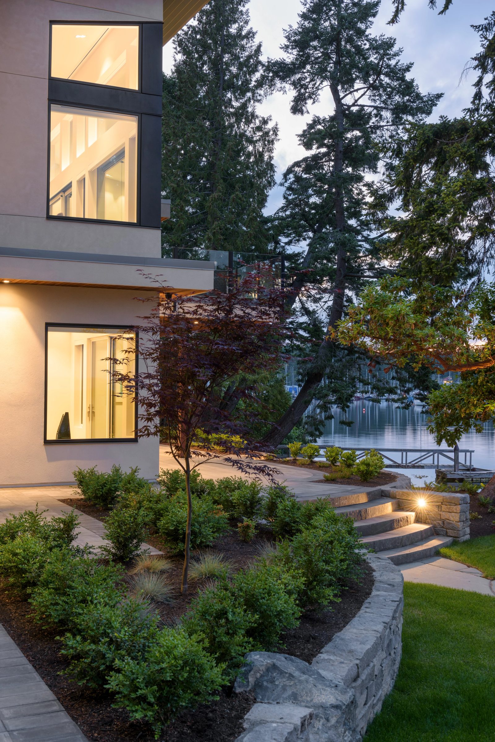Lit stone path descending past Japanese maples toward the water at dusk, with a marina beyond