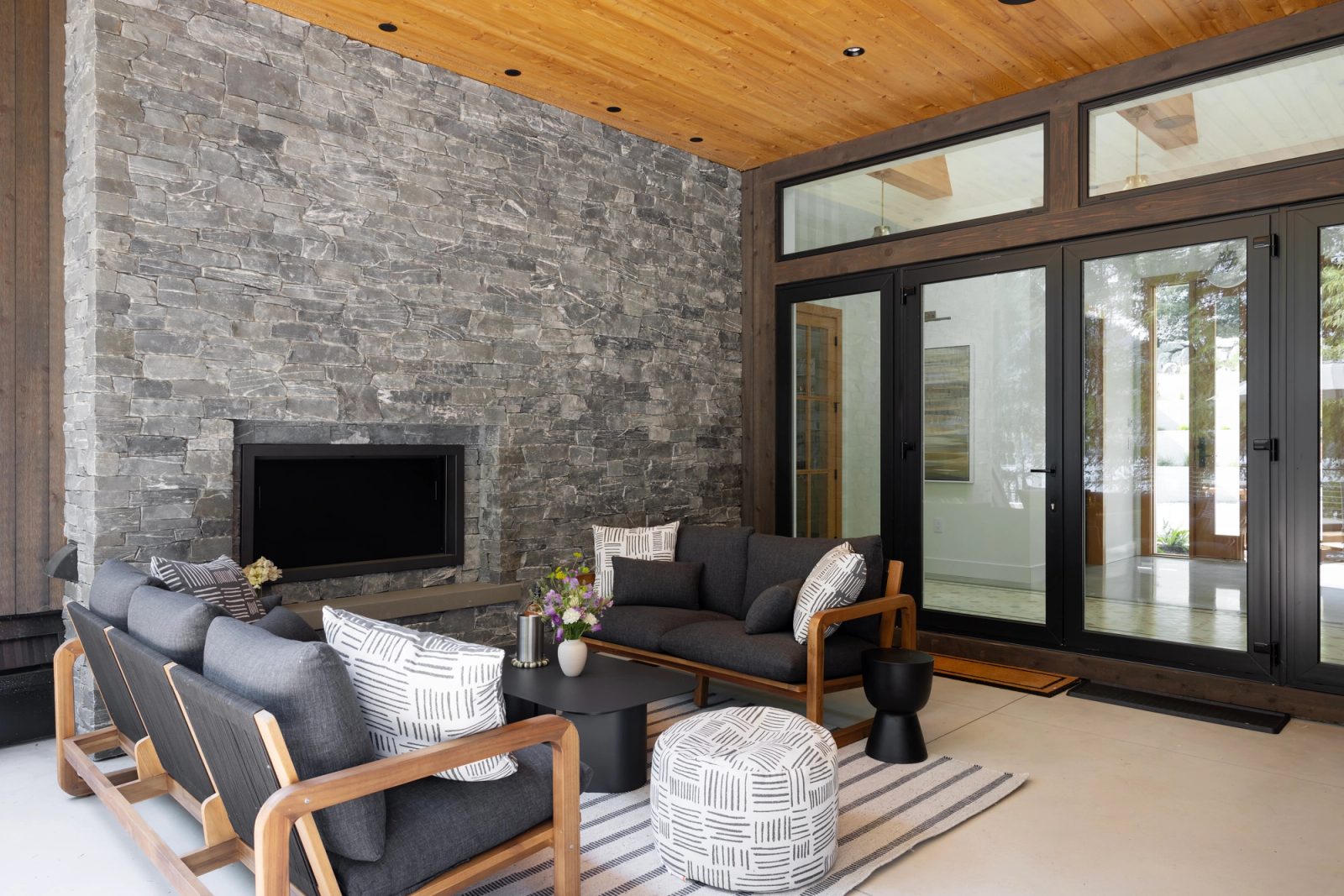 Covered outdoor room with a grey stone fireplace, cedar soffit and folding glass doors open to the terrace