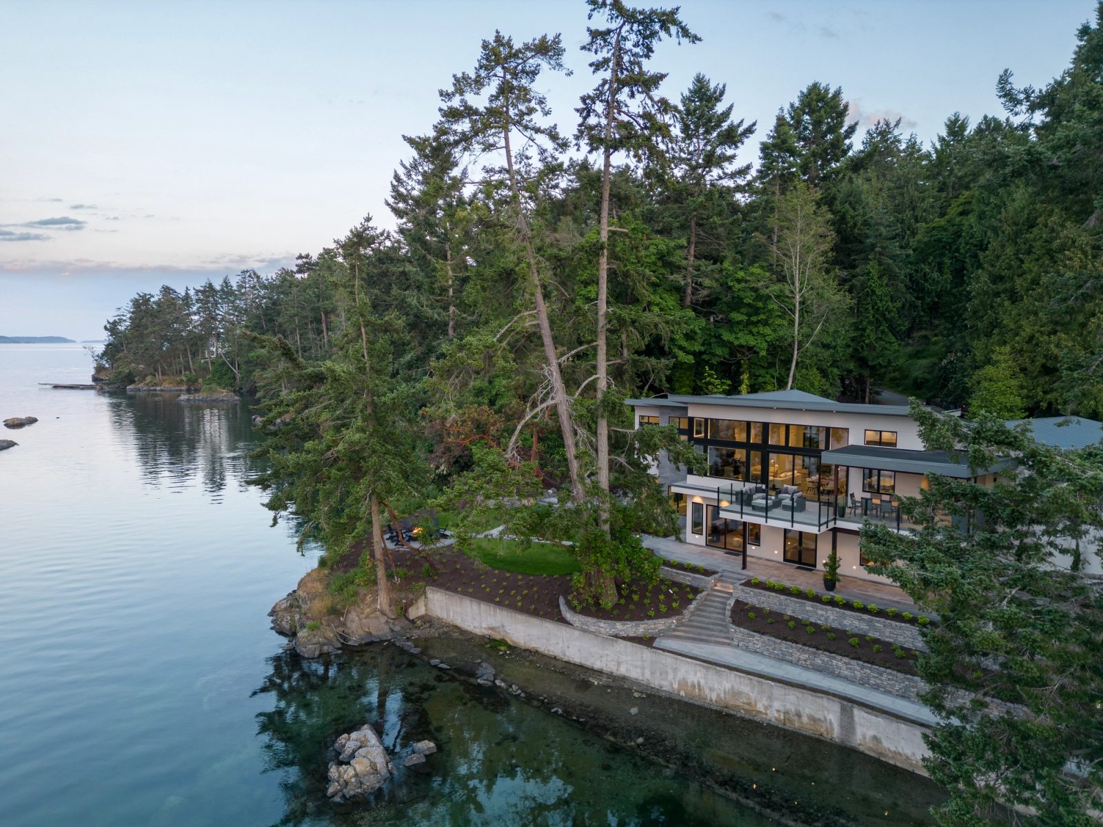 Aerial view at sunset of a waterfront house on a forested point, with open water to one side and a sheltered cove to the other