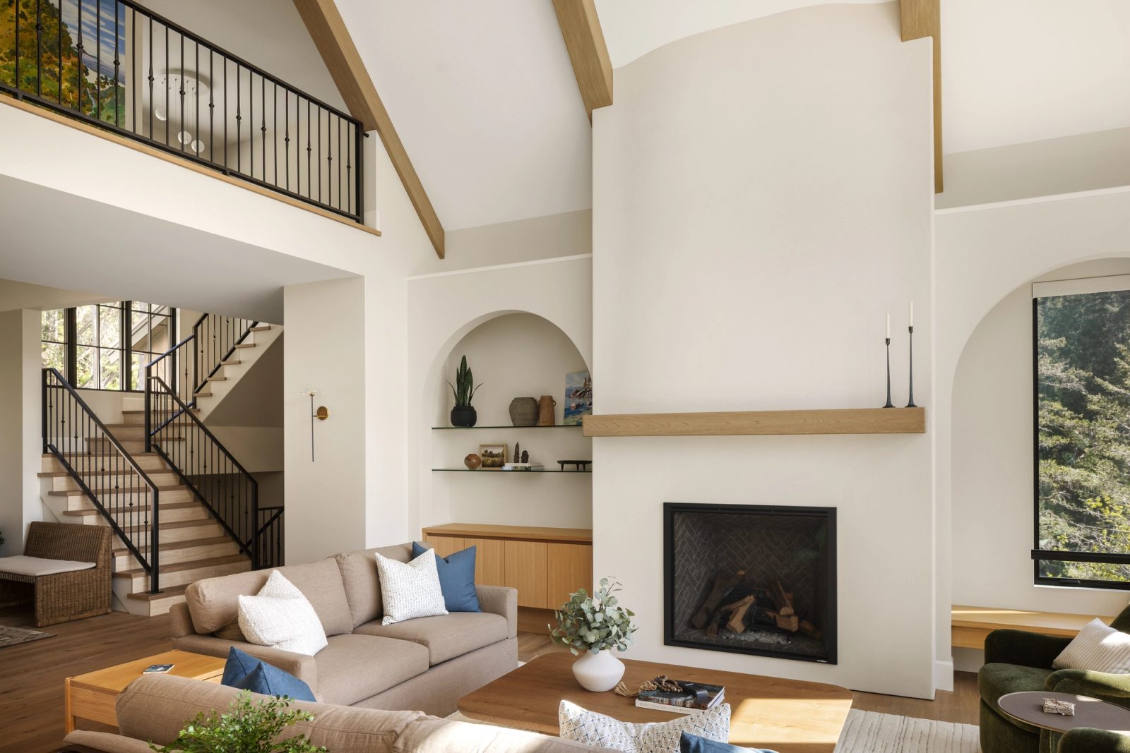 A double-height living room in pale plaster with an oak mantel and beamed vault, an open stair with a black iron rail beyond, and an arched niche between.