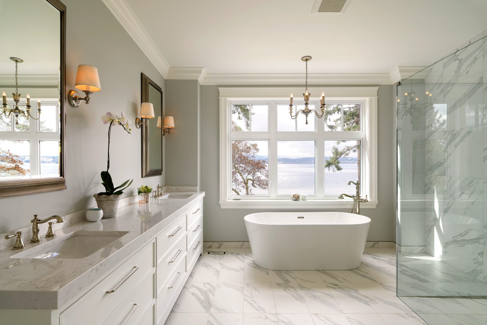 Freestanding tub beneath a triple window, a long double vanity in marble, and a glass shower enclosure alongside.