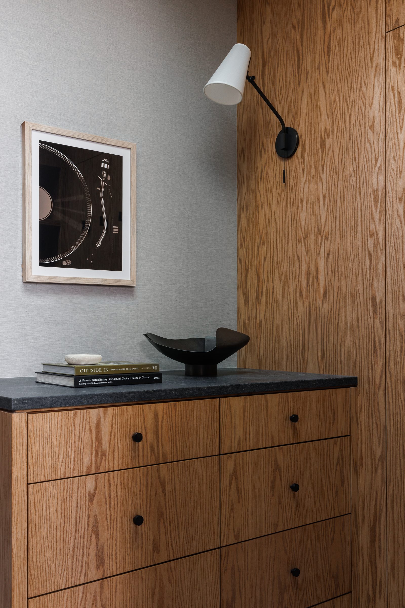 Right: a dresser in oak under a black stone top, a framed print on grasscloth and a black swing-arm light on the panel beside it.