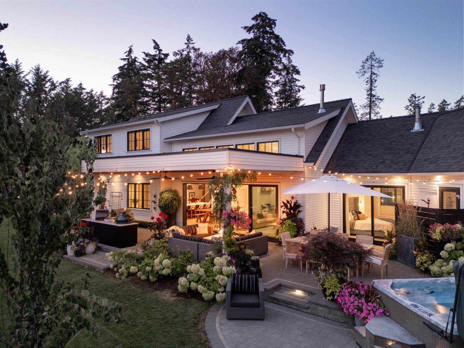 Covered patio at dusk strung with lights, with an outdoor kitchen, dining table, hot tub and sliding doors open to the living room and a bedroom