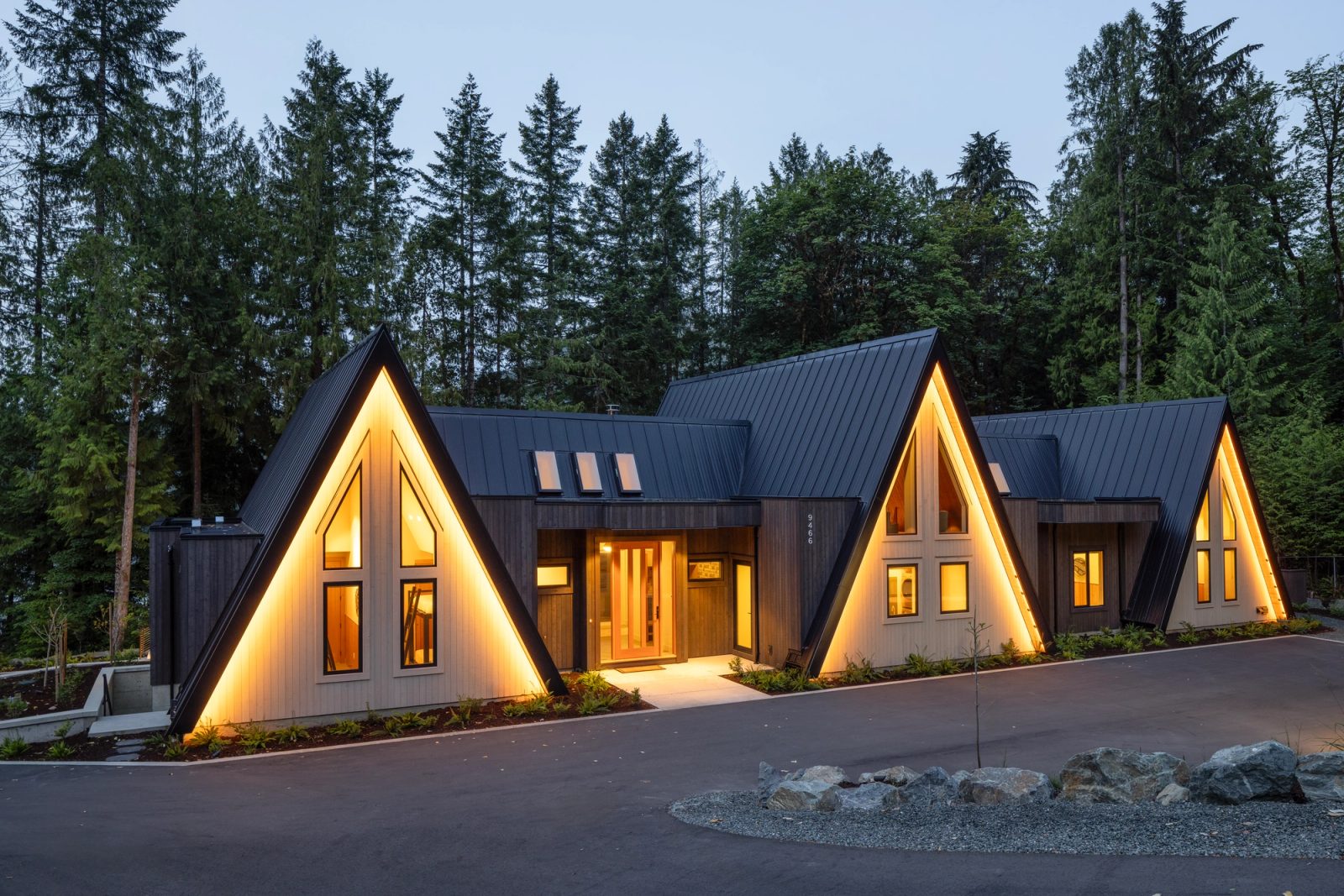 Three pale gables glowing at dusk above a black metal roof, firs behind
