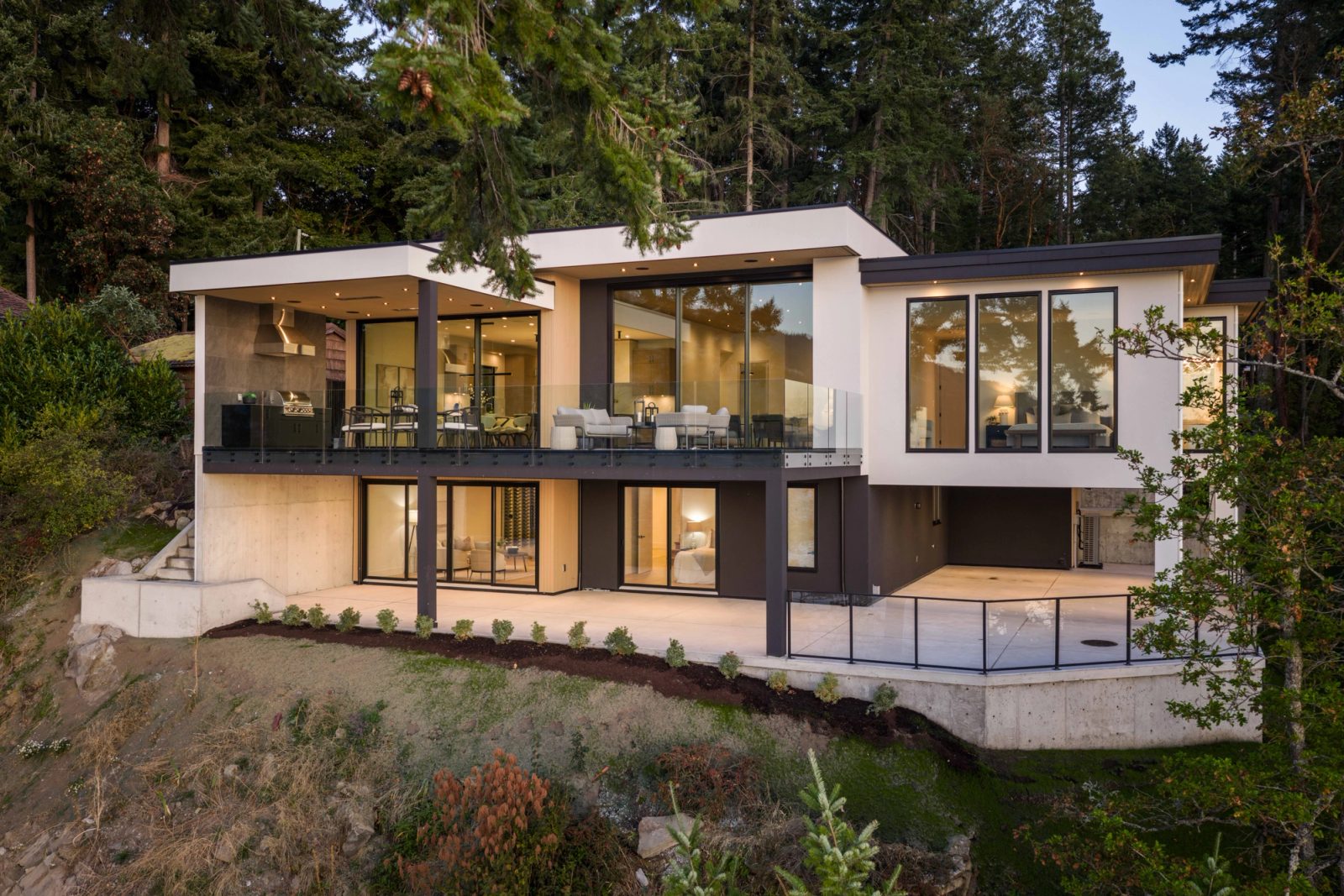 The whole house seen from below at dusk, both floors lit through full-height glass, standing on its concrete base among firs.