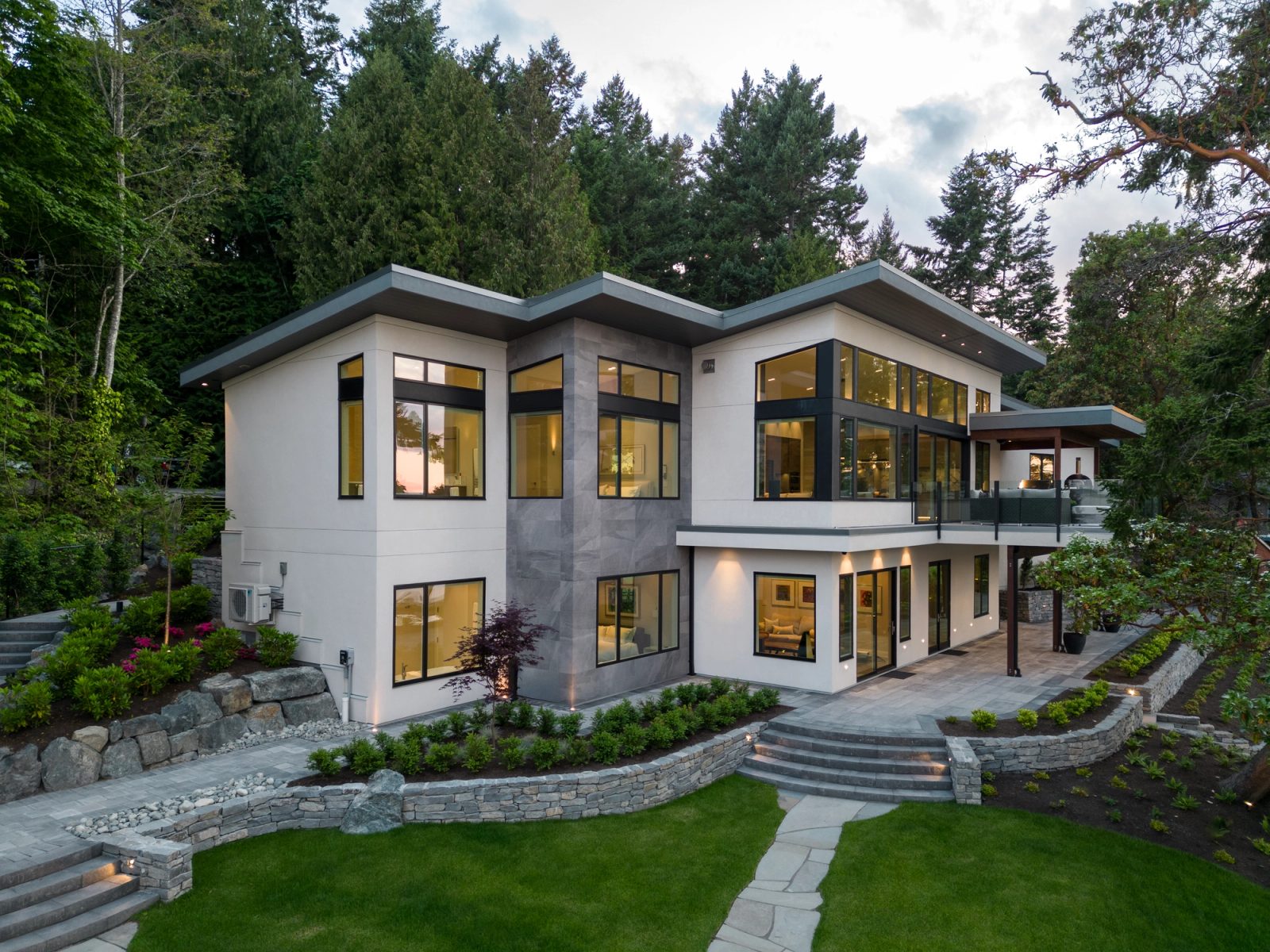 Waterfront elevation at dusk with a cantilevered deck, lit interiors and stone steps down to the lawn