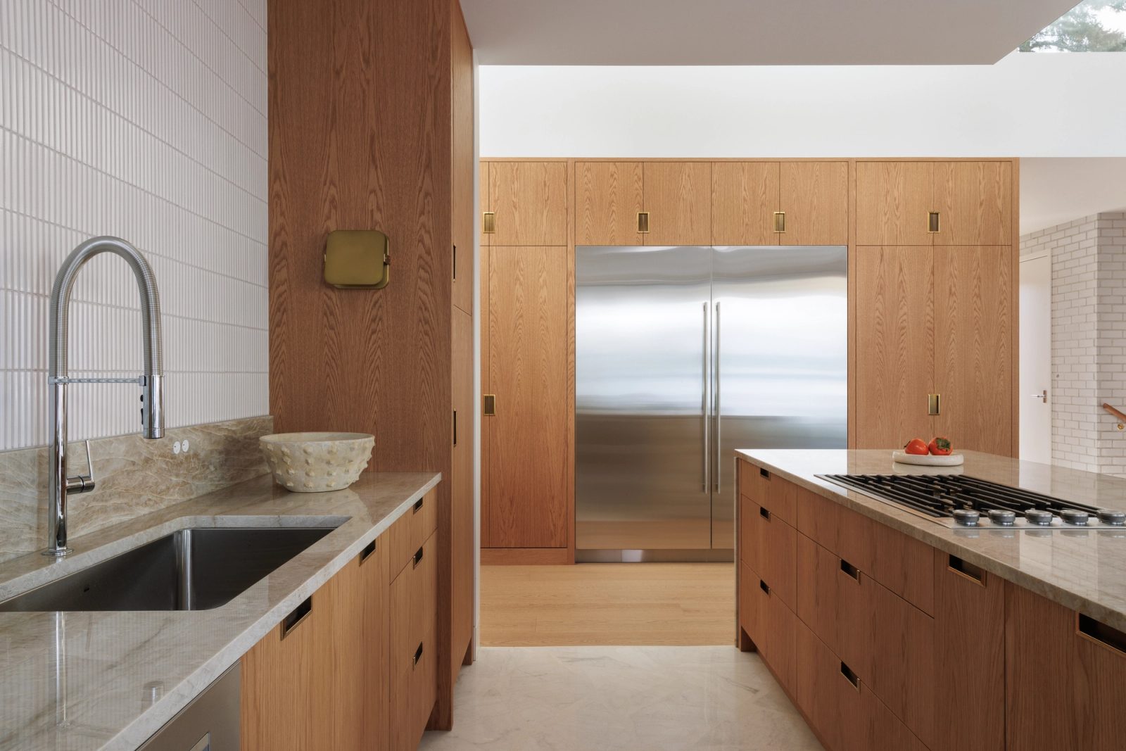 The kitchen looking through to a wall of full-height oak cabinetry with integrated refrigeration, a passage opening to white brick beyond.
