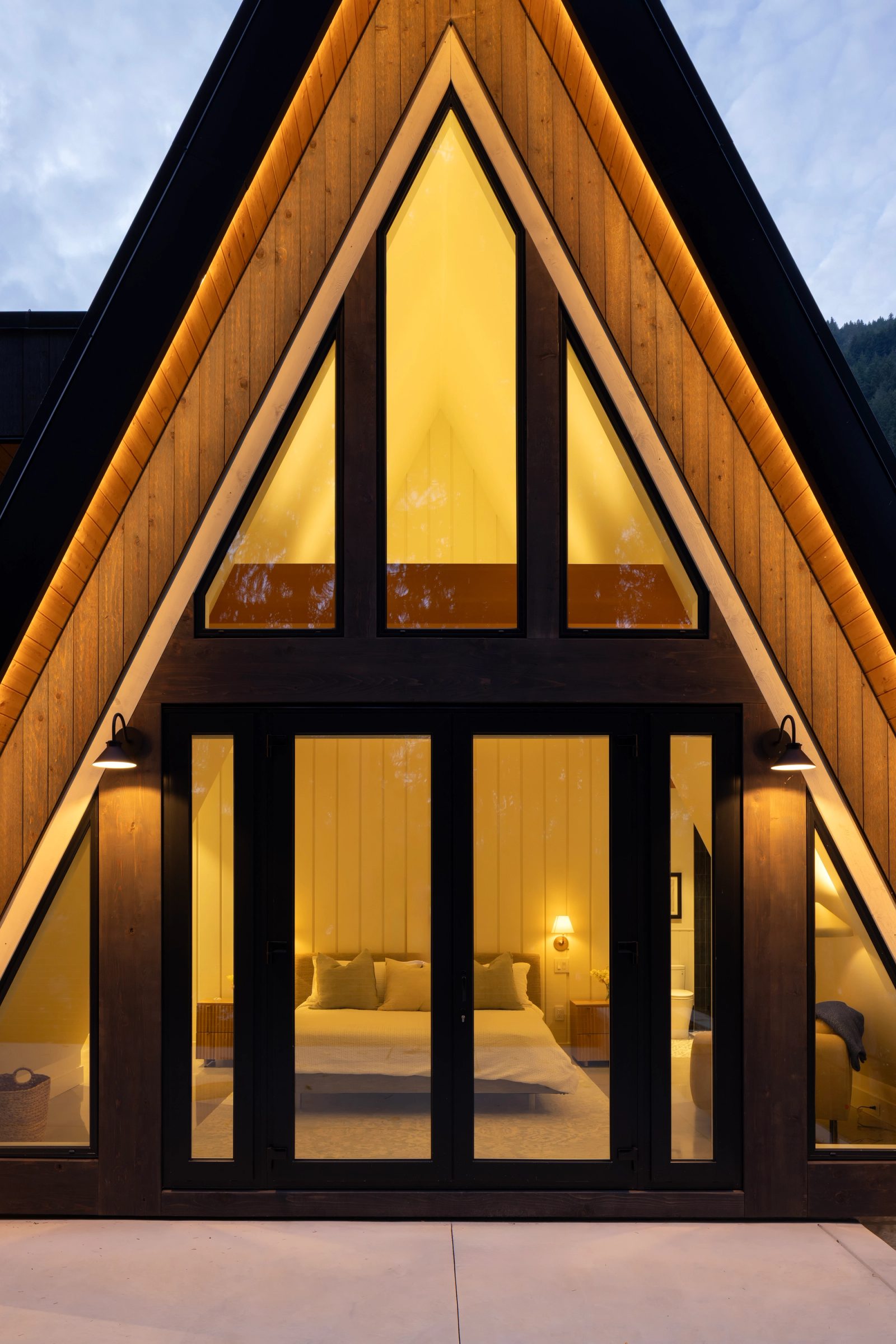 Lit A-frame gable framing a bedroom behind full-height glass at dusk