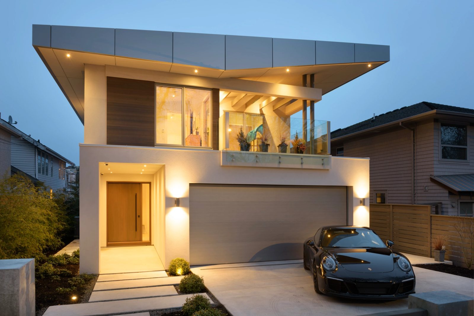 Entry recess lit warm beside a double garage door at dusk, a low car on the concrete apron and clipped shrubs along the walk.