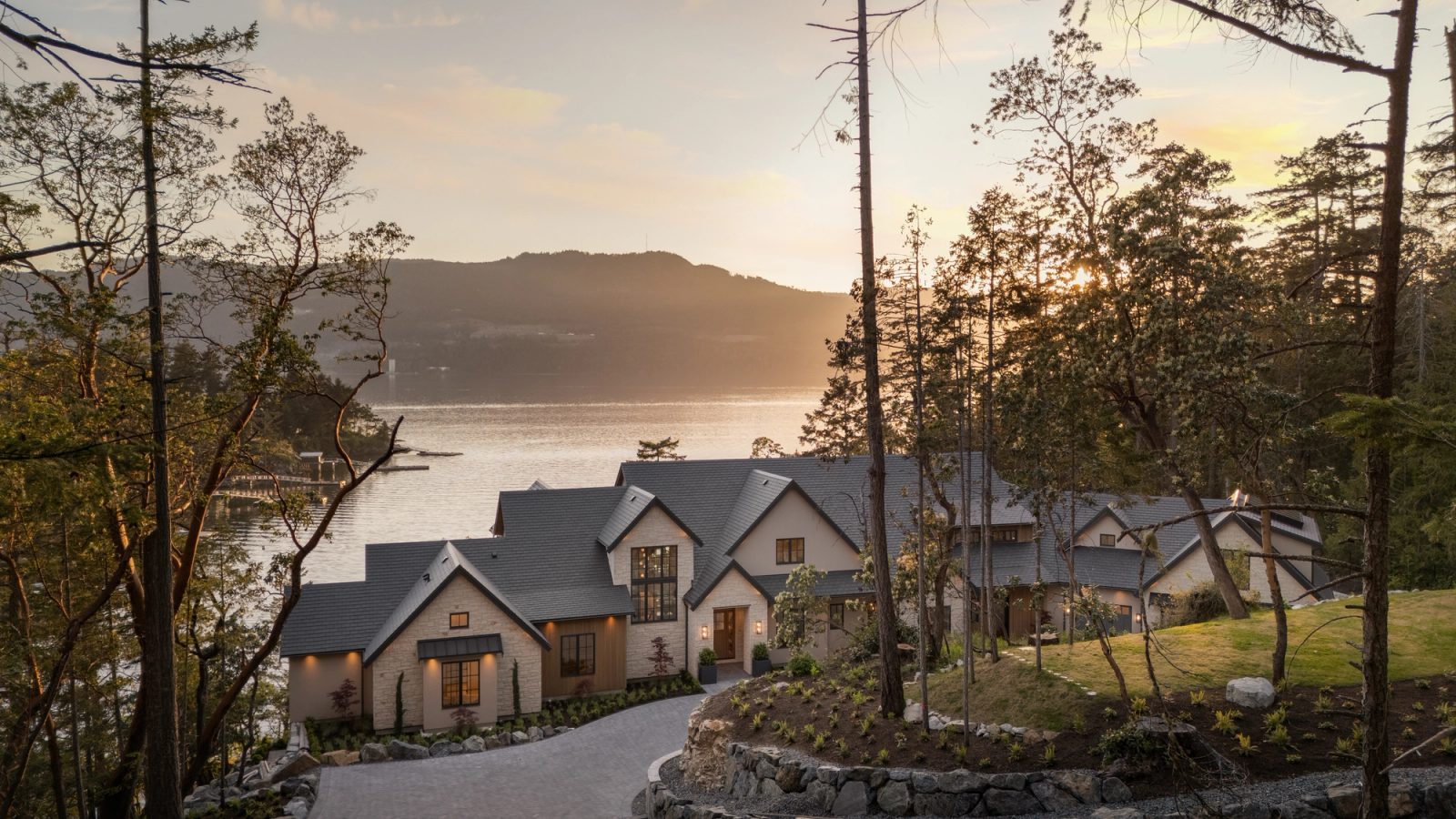 The house at last light from above the approach, its roofs stepping down through the trees, the inlet and the far shore going gold behind it.