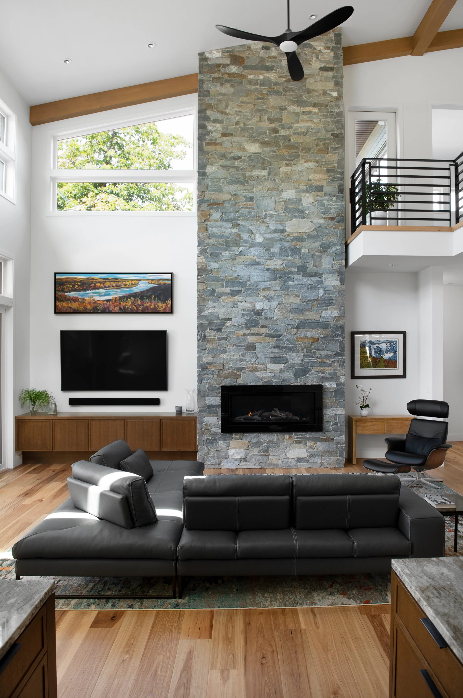 Two-storey living room with a full-height stacked stone chimney, beamed ceiling and a railed loft above