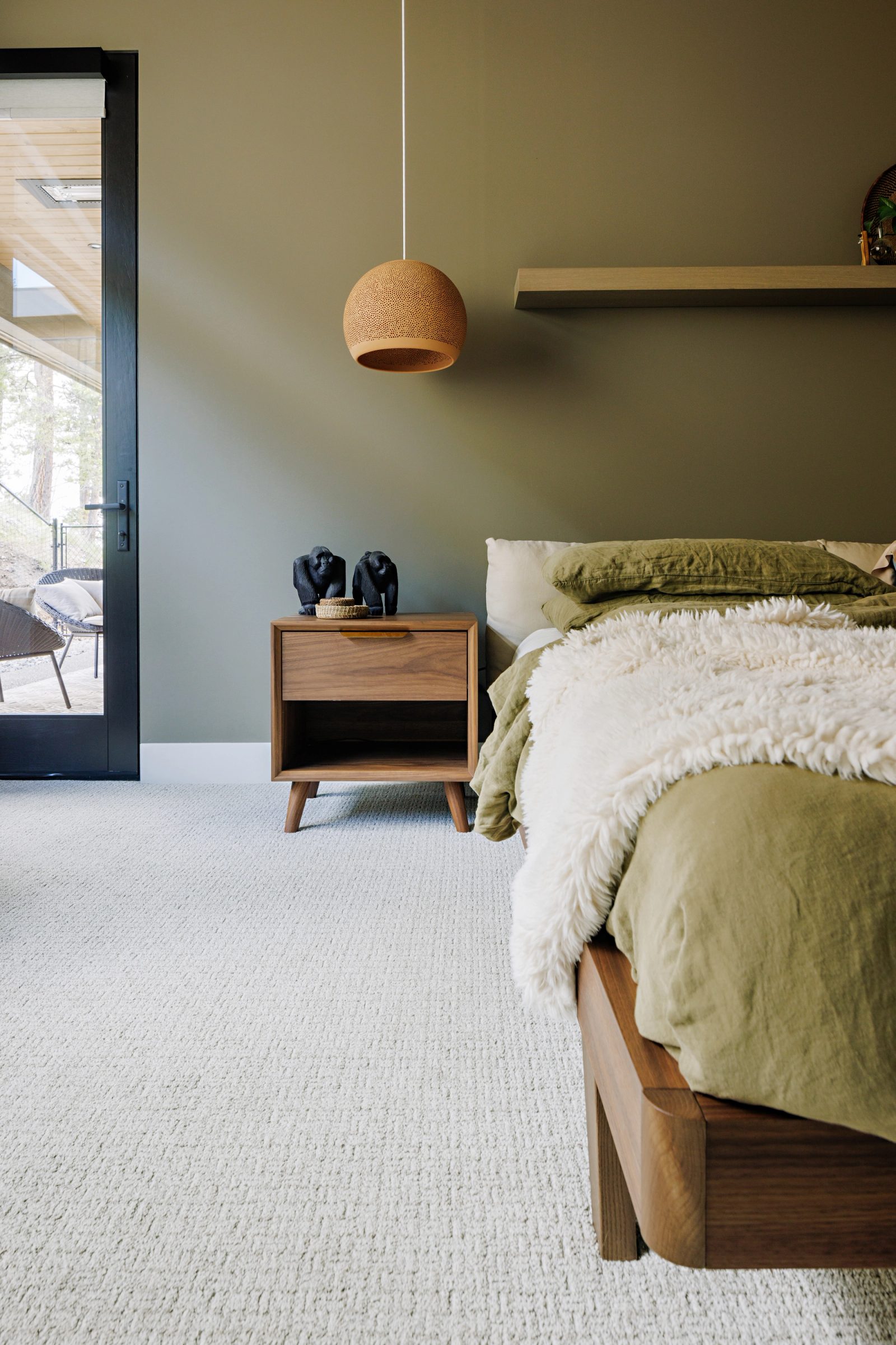 a bed dressed in olive linen against a deep olive wall, with a glazed door open to a covered terrace and pines beyond.