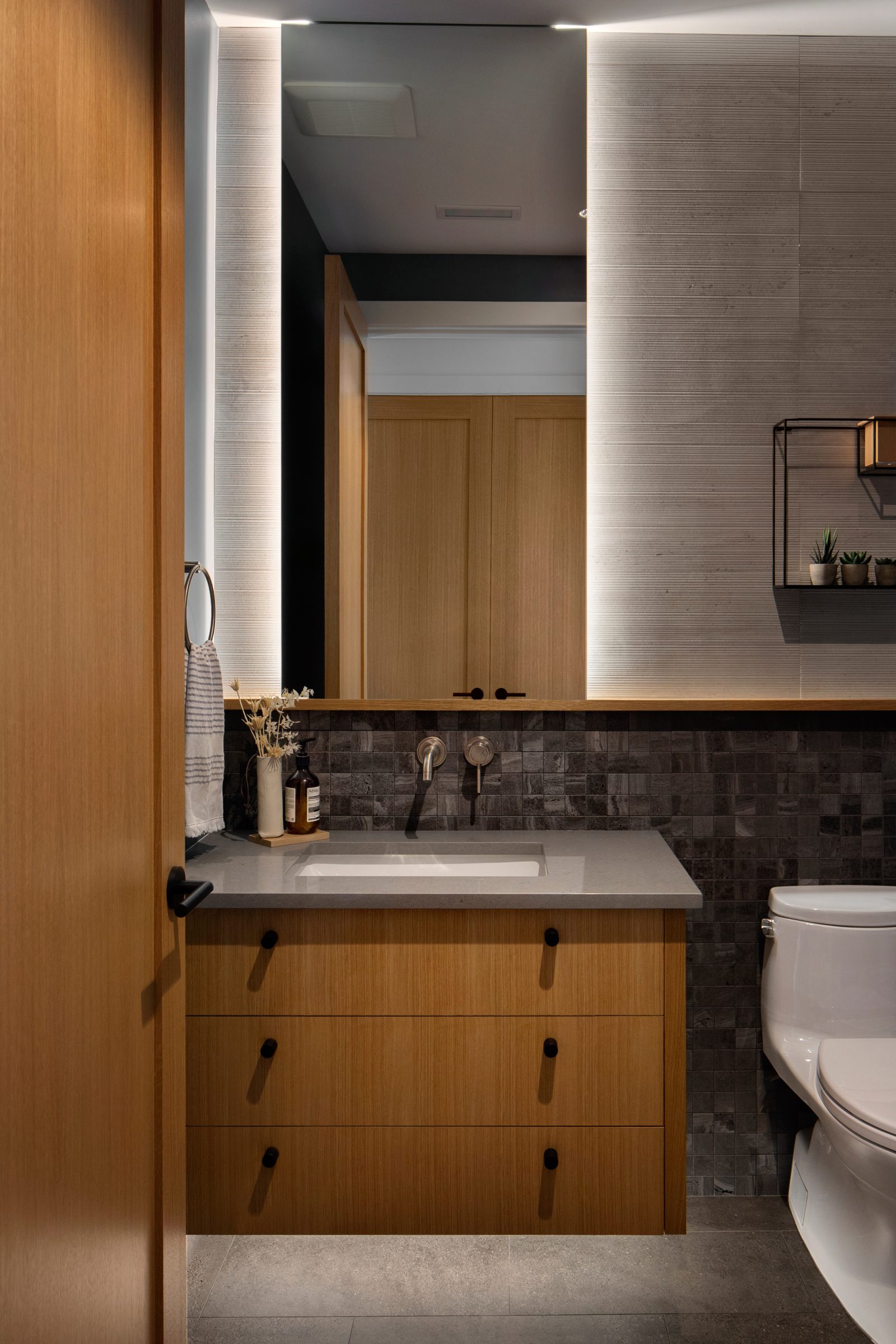 Powder room in dark stone mosaic with a backlit mirror and oak vanity