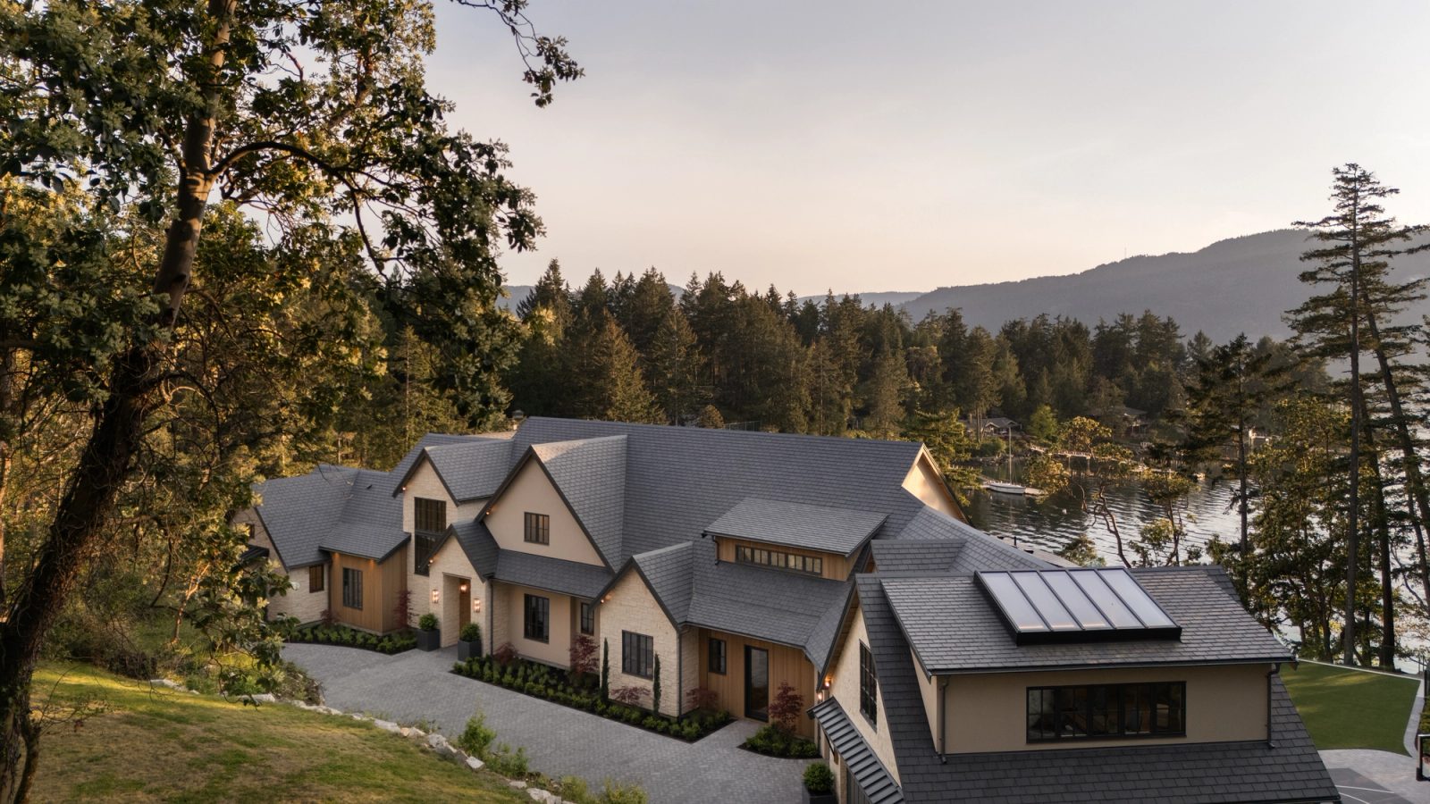 The roofscape at dusk from higher still, the entry court lit below, the cove and a moored sailboat past the trees.