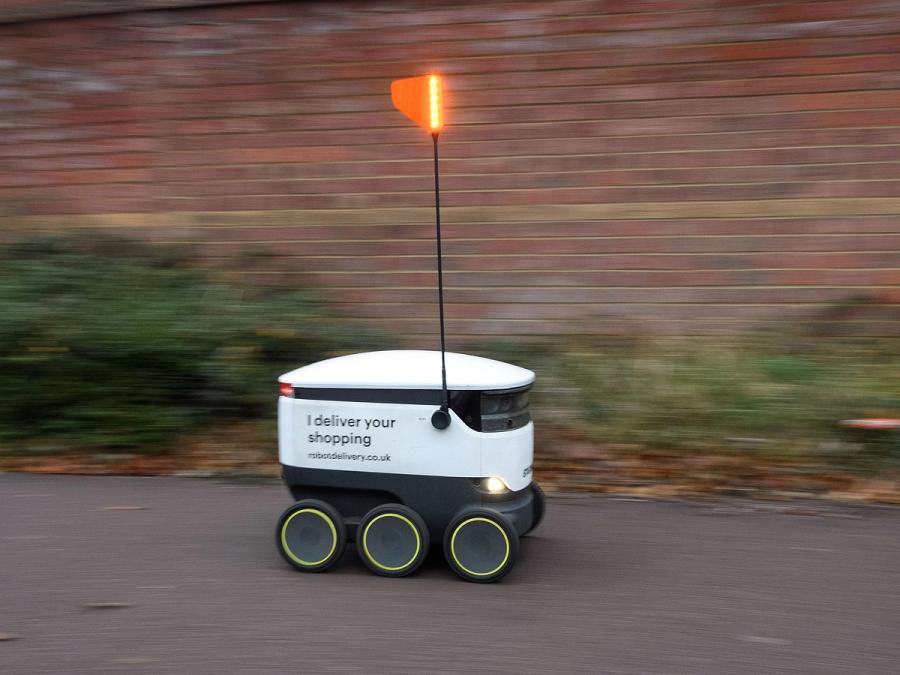 A white boxy mobile robot on six wheels with a narrow antenna with an orange flag on top speeds down a street.