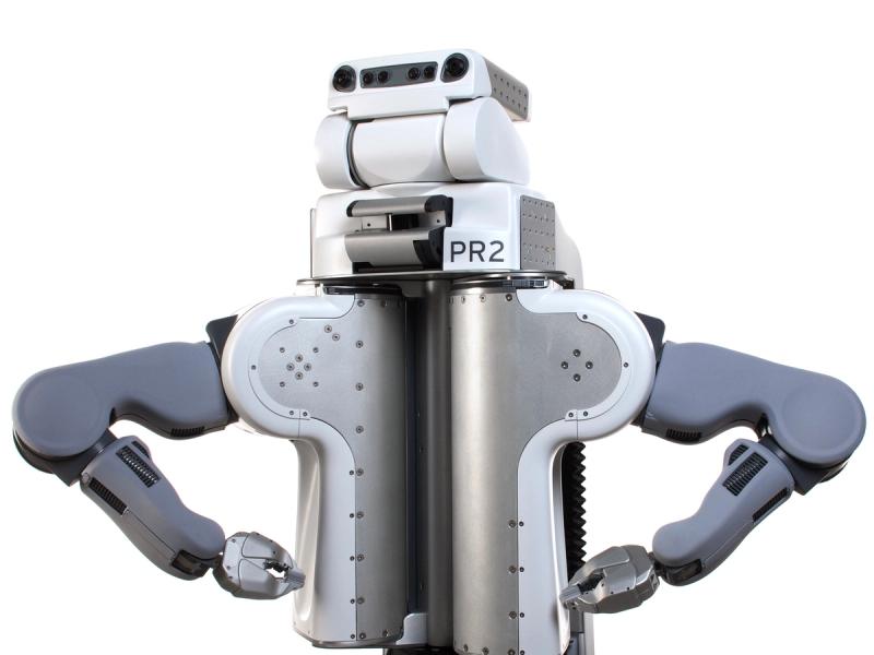 A white plastic and metal torso holds three stacked sections, the bottom which says PR2 and the top of which has two eye cameras and a black bar with sensors. The robots arms, with gripper end effectors, are posed on it's hips.