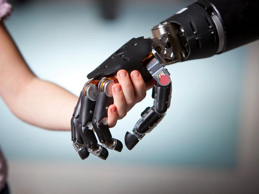 A child's hand intertwines with a black robotic hand with five jointed fingers mimicking a human hand.