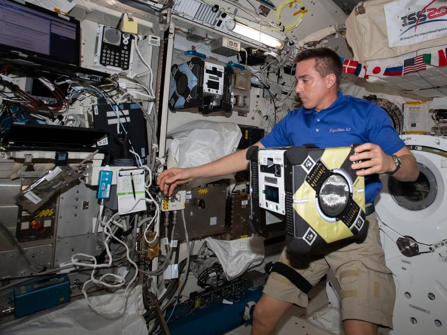 An astronaut has one hand on a floating Astrobee as he reaches towards something on the International Space Station, which is full of electronics.