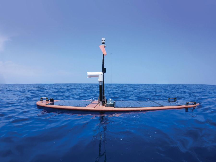Side view of copper board-like shape covered in solar panels that floats on the open water and has poles with cameras and sensors rising up.
