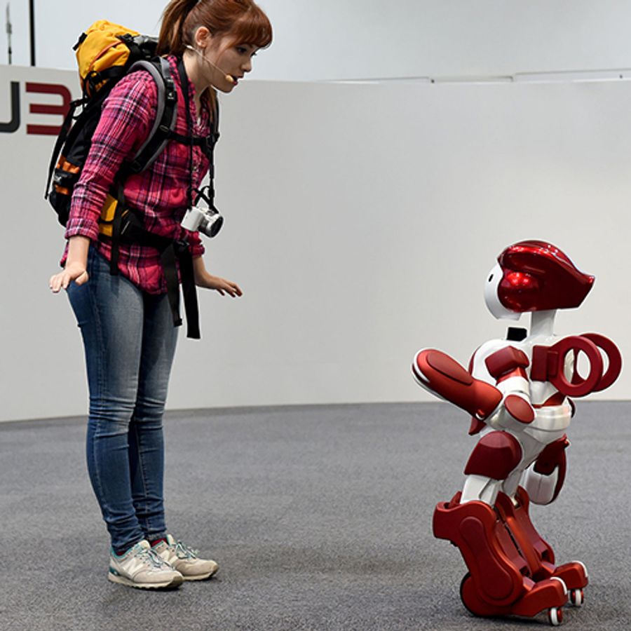 A person with a backpack addresses Emiew, a red and white humanoid who is half the size of the human.