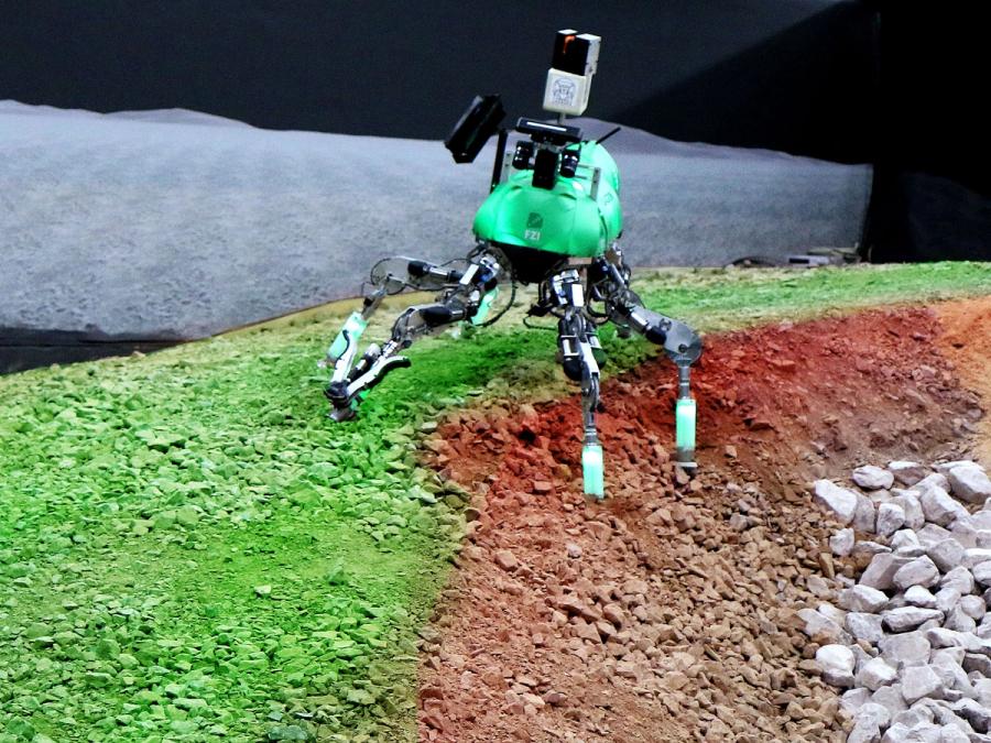 The robots six legs are spread between a level and rocky angled terrain.