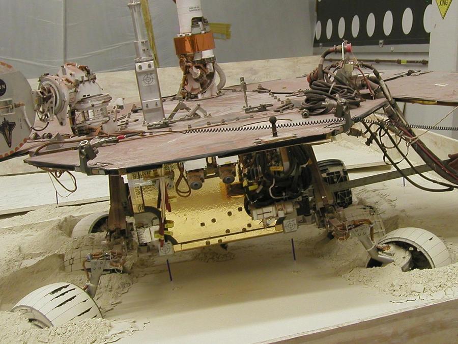 A version of the rover is seen with its wheels partially buried in beige sand, in a lab.