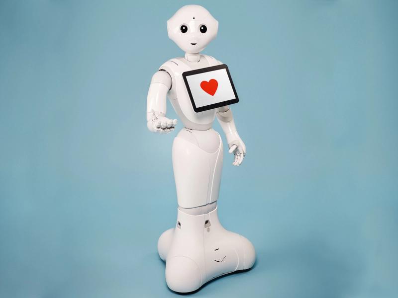 A friendly, gesturing, white humanoid robot with a mobile base, simple smiling face, and a tablet on its chest with a red heart symbol.