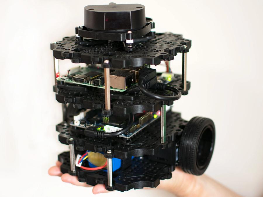 A modular black mobile robot with two rubber wheels and a scalable three layered structure of 3D-printed plates, each of which houses electronics and sensors. The whole robot fits in the palm of a hand.