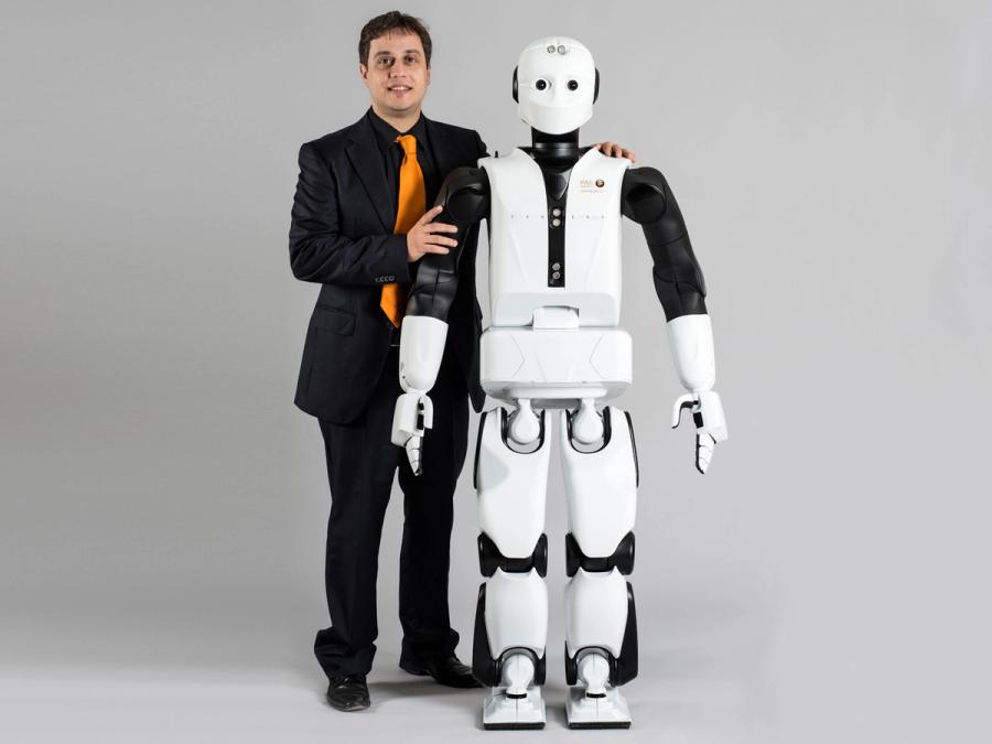 A man in a black suit and orange tie puts his arm around a white and black humanoid robot that is only slightly shorter than him.
