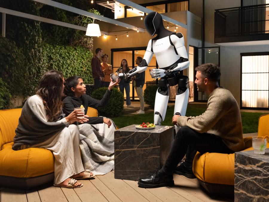 Tesla's Optimus, a silver and black bipedal humanoid robot, hands out drinks to seated humans at a party.