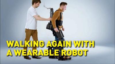 A man wearing the Atalante exoskeleton, which consists of a pair of robotic legs attached to the user's own legs, takes a step, assisted by another man who's holding on to the device, with superimposed text that reads, Walking again with a wearable robot.