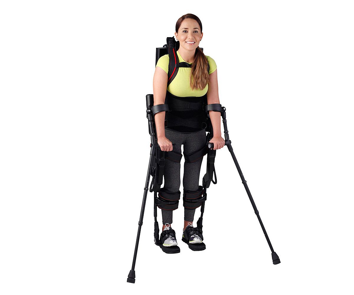 A woman in a yellow shirt wears a black exoskeleton backpack suit, and is supported by crutches attached to her arms.
