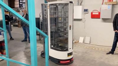 A tall mobile robot, with metal racks encased by a transparent plastic shell, drives inside a warehouse.
