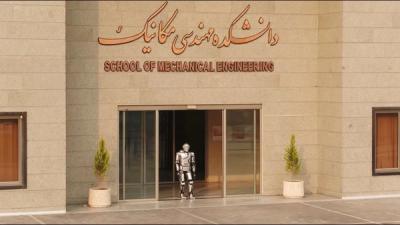 Surena 4, a silver adult-size humanoid robot, walks out the front glass sliding doors of a beige building where the School of Mechanical Engineering is located.
