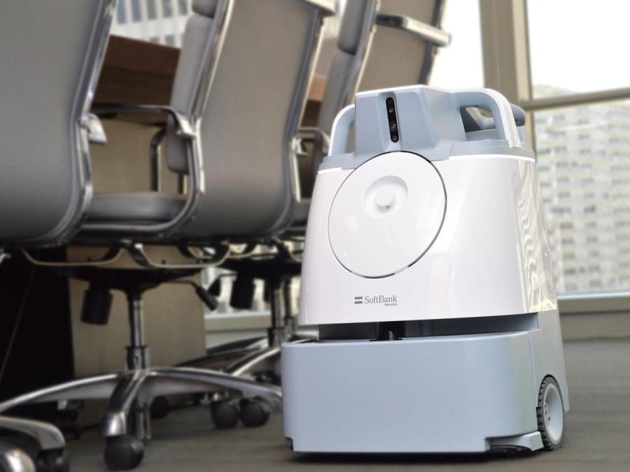 A white and gray boxy shaped robotic vacuum on a wheeled base rolls alongside office chairs in a conference room.