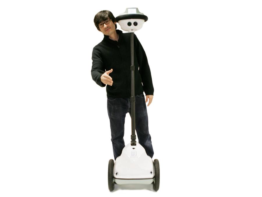A smiling man reaches a hand out from behind a telepresence robot. It has a white base with two black wheels, a simple center composed of a tall, thin adjustable black pole, and a white head with camera eyes and a display on top.