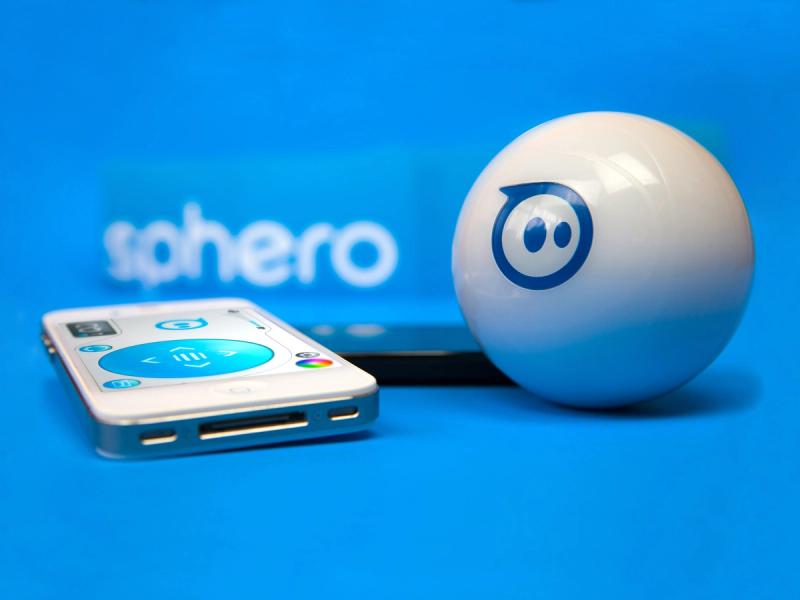 A white ball with a logo on it like a blue circle with two eyes next to a smartphone displaying controls.