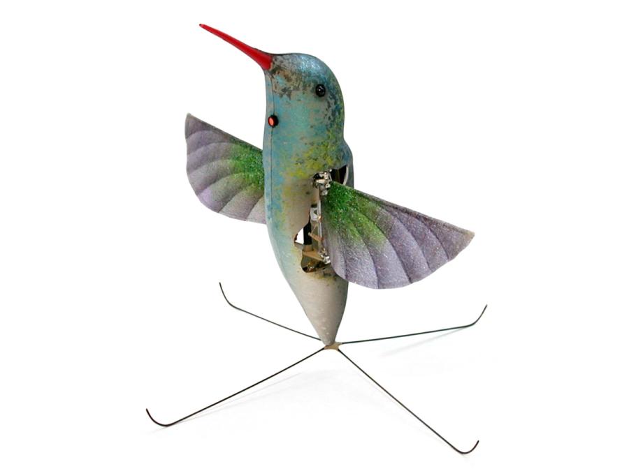 A robot that looks like a hummingbird including wings and long, thin, orange bill stands on a four pronged base.