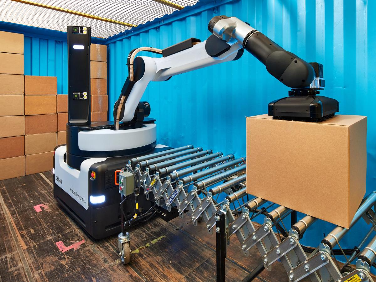 Boston Dynamics' Stretch robot has a large black and grey mobile base which supports an articulated arm with a suction pad that picks a cardboard box off a conveyor. A stack of boxes is in the background.