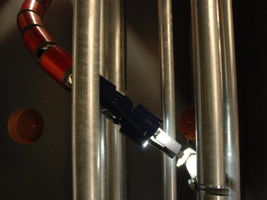 A red, snake-like segmented robot moves between pipes with a gripper hand reaching towards the pipe in front of it.